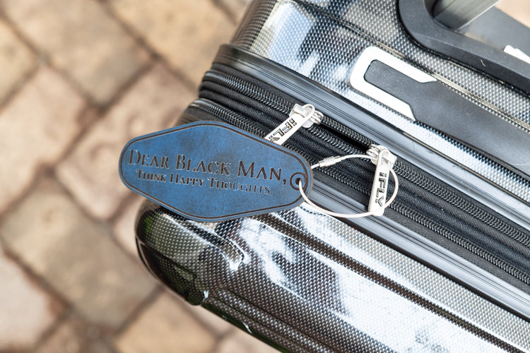 Blue leather tag reading Dear Black Man, Think Happy Thoughts attached to a black suitcase